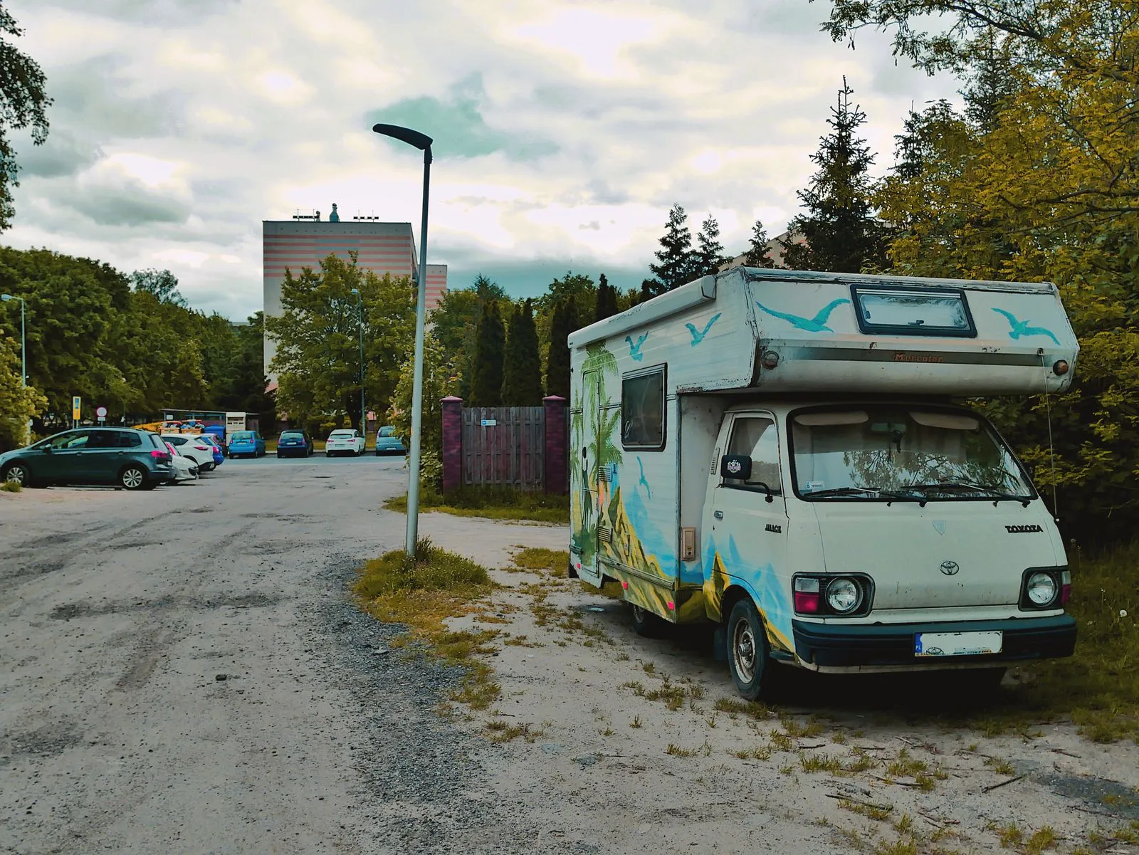 An old painted motorhome parked on a gravel lot at the edge of a city parking area