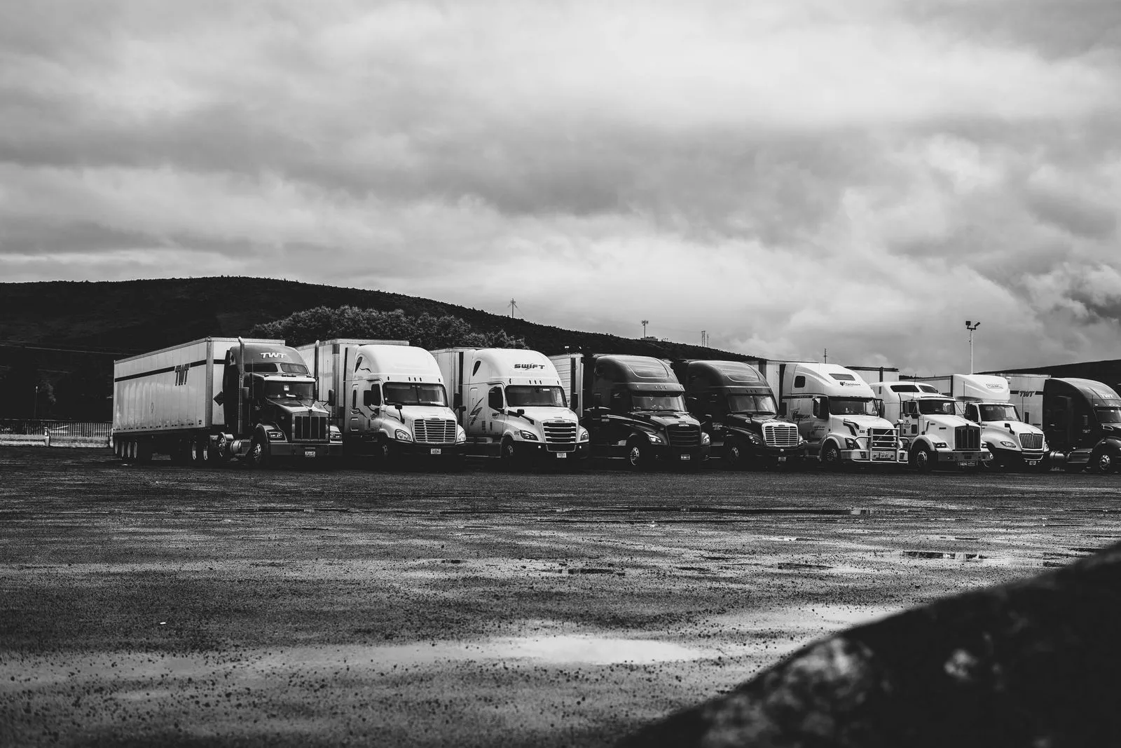 A row of semi tractors parked on a wet gravel lot under a cloudy sky, in black and white