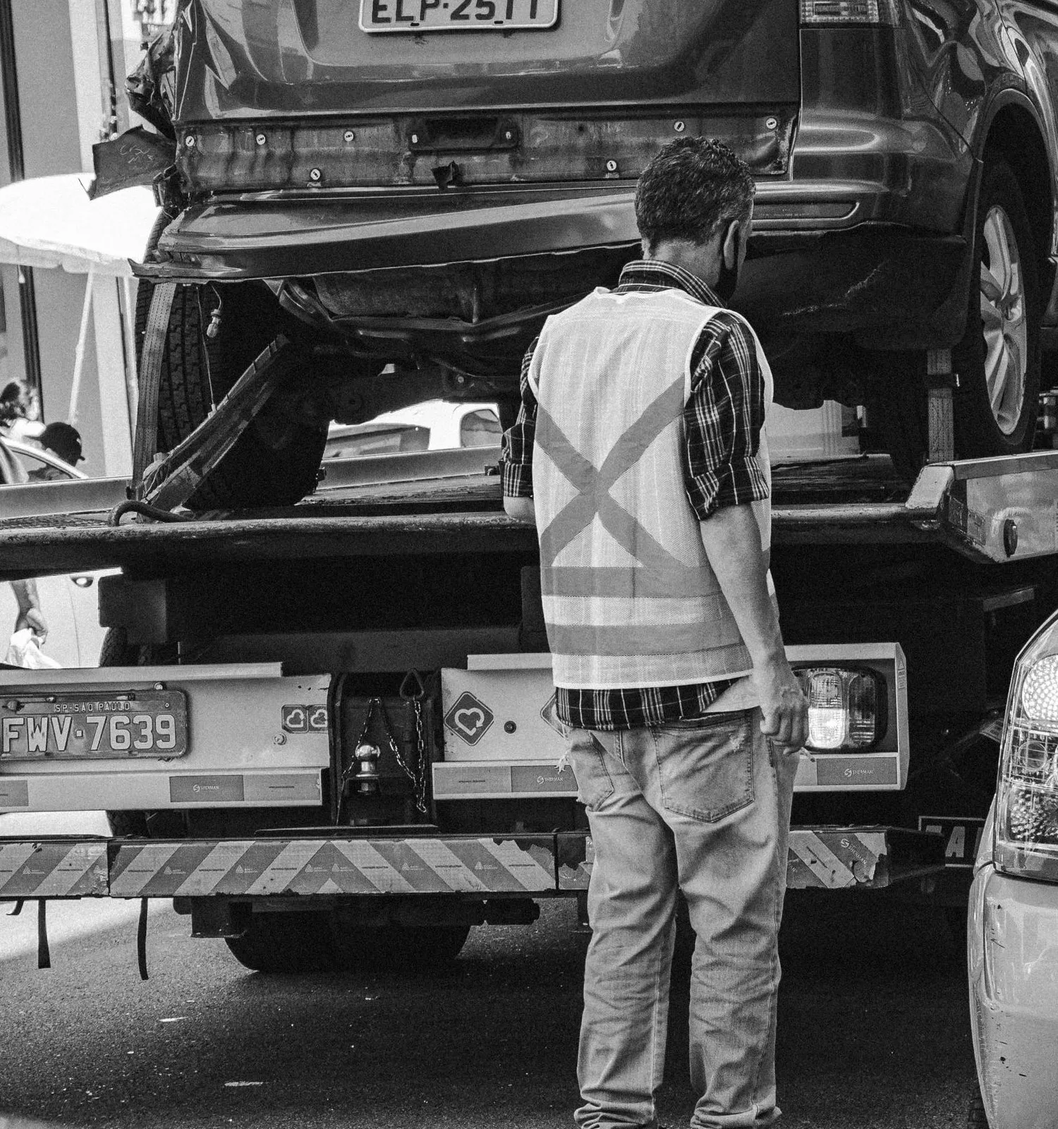A worker in a safety vest stands behind a flatbed tow truck carrying a damaged SUV on a city street