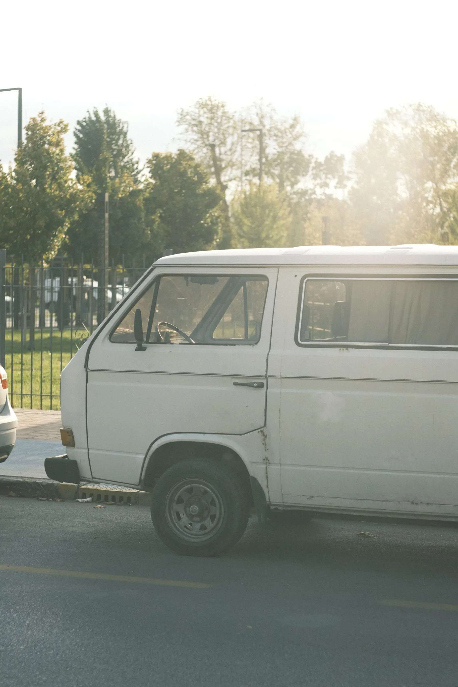 An old white van parked at the curb on a sunny street lined with trees